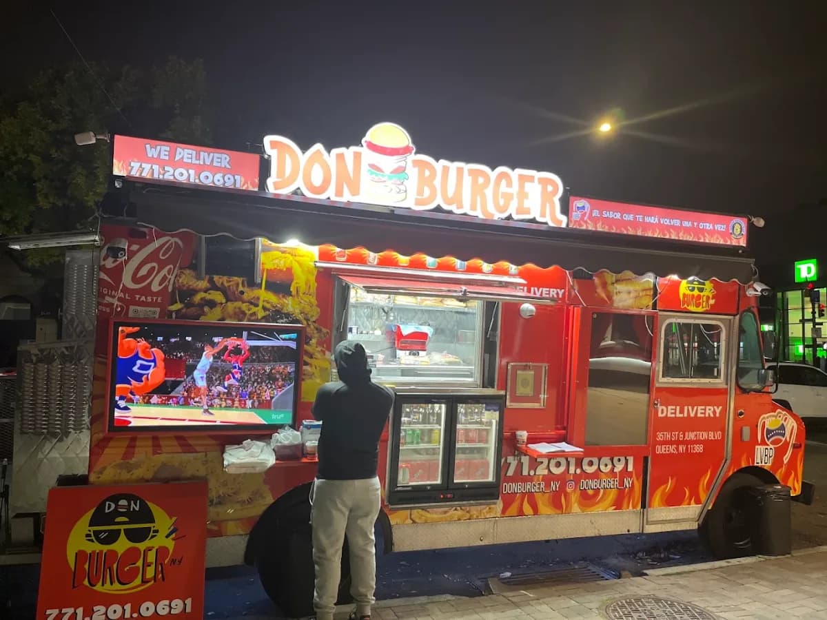 The red Don Burger Venezuelan food truck lit up at night with its glowing burger sign, a flat screen TV mounted on the side and a customer ordering at the window