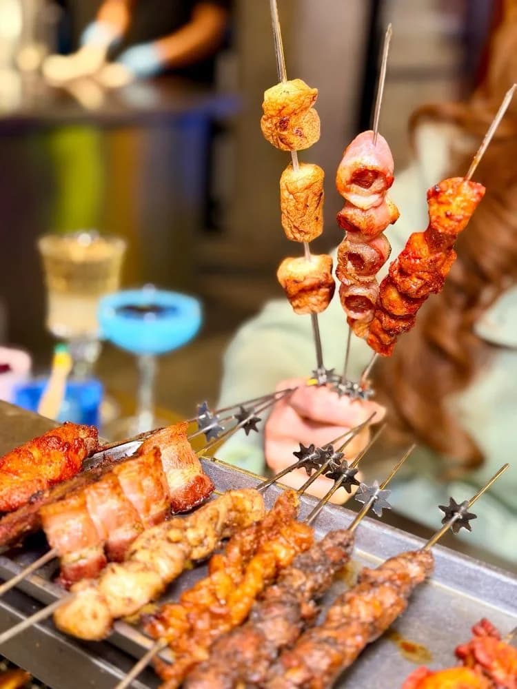 Skewers of pork belly, chicken and beef turning on the automated grill at GanBlaze Chinese BBQ in Flushing, with a blue cocktail in the background