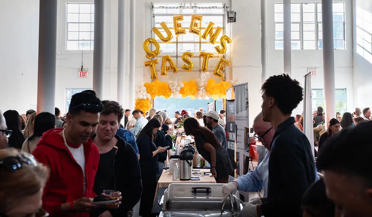 A packed Queens Taste tasting event in a bright loft space, gold balloon letters spelling QUEENS TASTE above a crowd browsing tables of food from local vendors