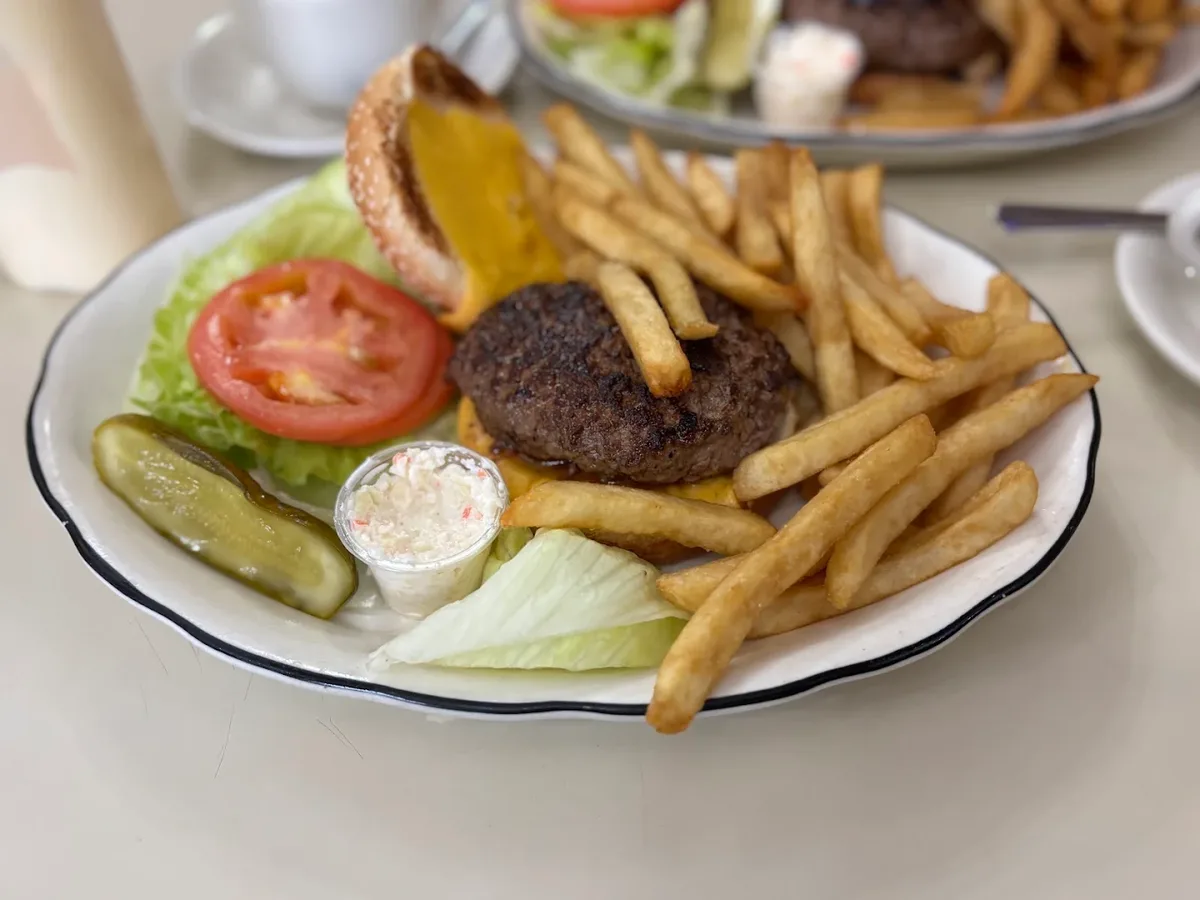 A classic diner cheeseburger at Joe Junior in Gramercy, served on a white plate with fries, lettuce, tomato, a pickle spear and a cup of coleslaw