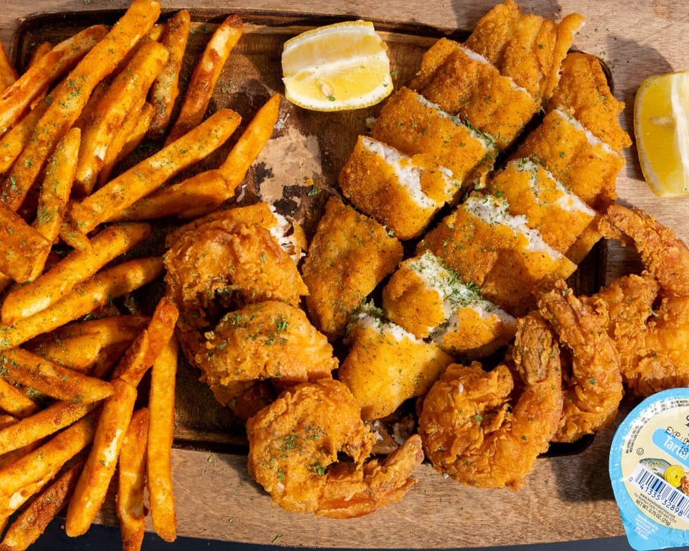 A fried fish and shrimp platter with seasoned fries, lemon wedges, and a cup of tartar sauce on a wooden board