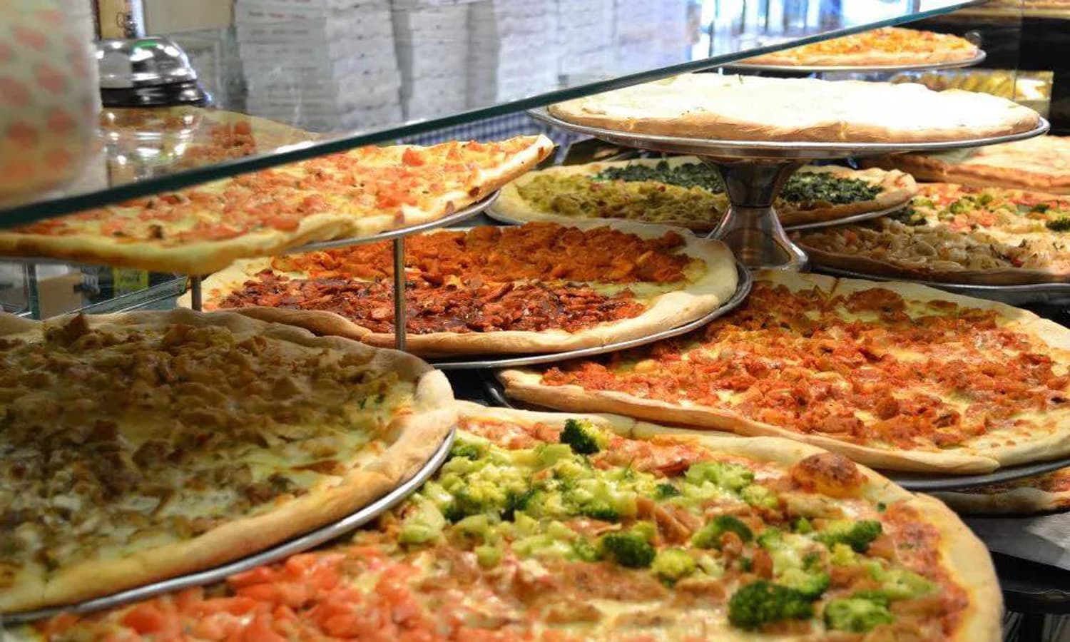 A pizza shop counter lined with fresh whole pies under glass, including chicken, broccoli, spinach, and buffalo chicken slices