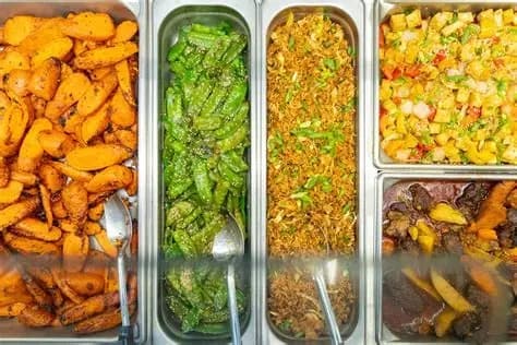 Steam trays of West Indian food at a supermarket hot bar with candied carrots, greens, rice, and stewed meats