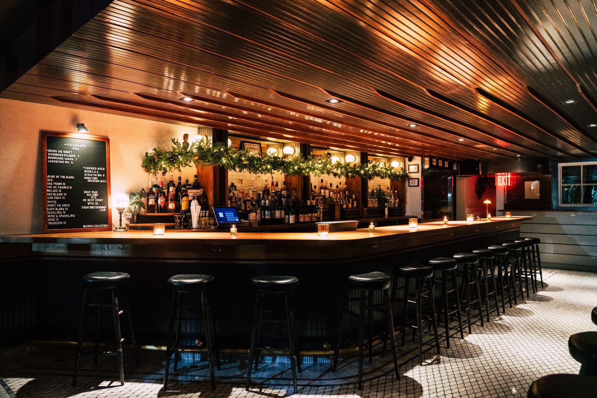 The long wooden bar at Charlie's in Astoria, lit warm under a slatted wood ceiling with a garland of string lights over the back bar and a row of black stools