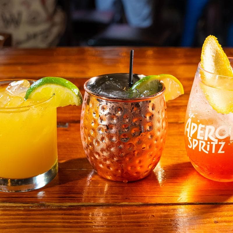 Three cocktails lined up on the bar at Murphy's in Astoria: a margarita on the rocks, a copper mug Moscow mule, and an Aperol Spritz with an orange twist