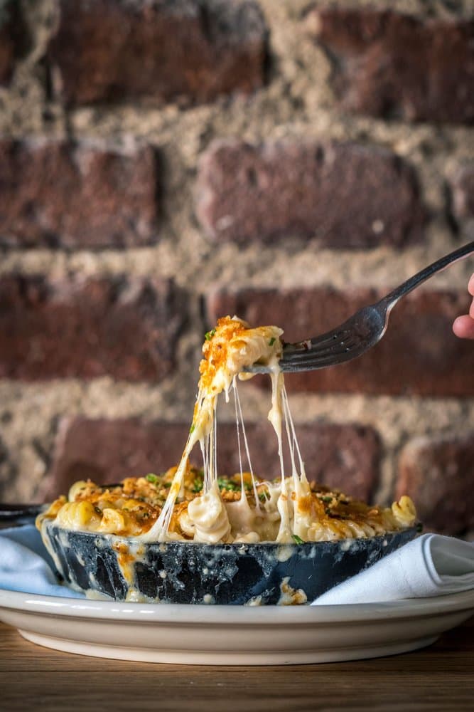 A fork pulling a stretchy, cheesy bite of baked mac and cheese out of a black skillet in front of a brick wall at Sweet Afton in Astoria