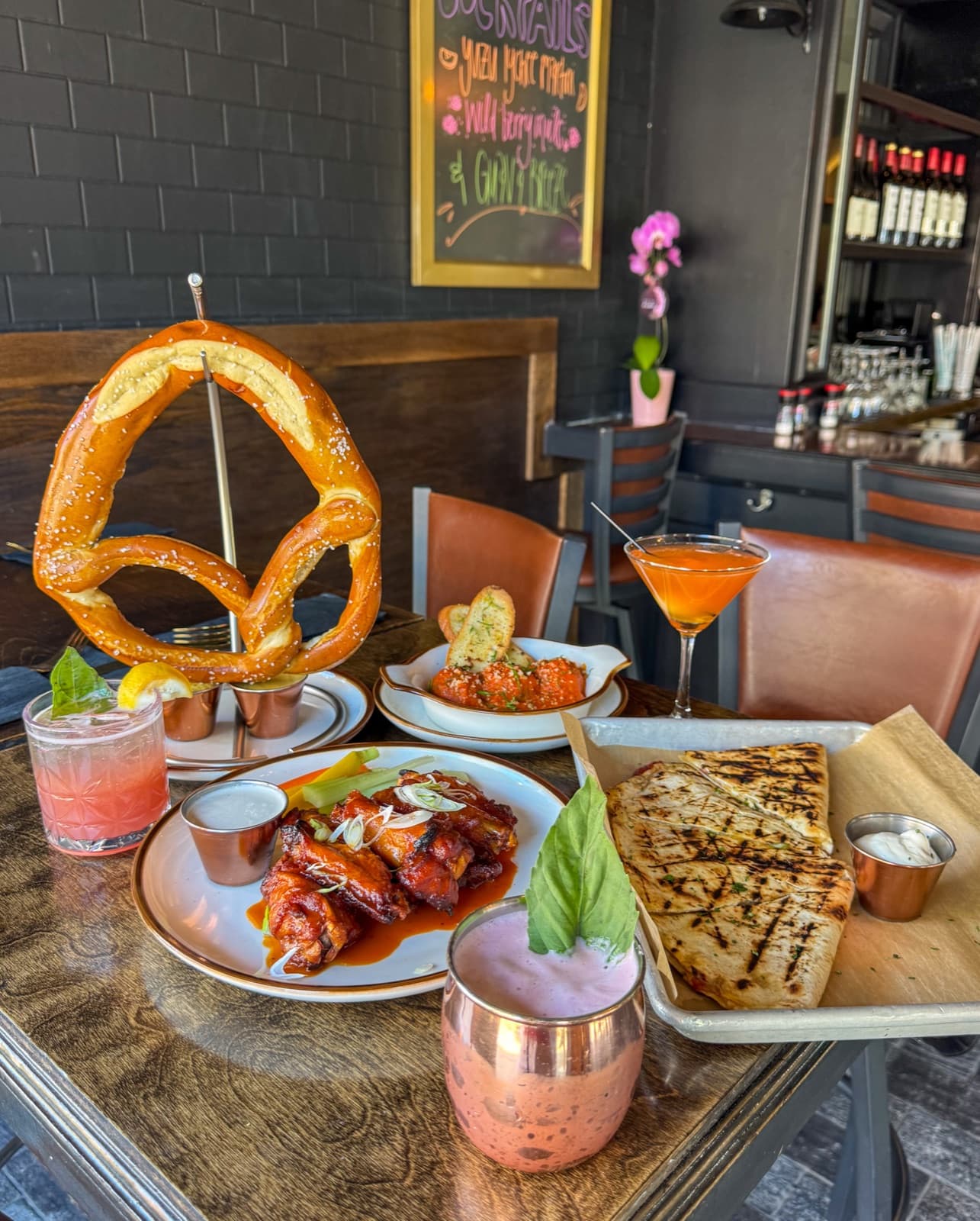 A spread at The Corner House in Astoria with a giant hanging pretzel, saucy wings, meatballs, a grilled flatbread, and cocktails